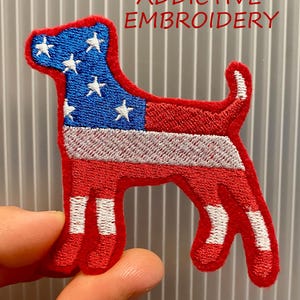 May include: Embroidered patch in the shape of a dog, featuring the American flag design. The dog's head is blue with white stars, and the body is red and white striped. The text "ADDICTIVE EMBROIDERY" is in red.