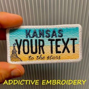 Könnte beinhalten: Gestickter Aufnäher mit einem Design, das einem Nummernschild aus Kansas ähnelt. Der Aufnäher zeigt das Wort "KANSAS" in Türkis, "YOUR TEXT" in Schwarz und "to the stars" in Schwarz. Der Hintergrund hat ein türkis-gelbes Farbschema. Der Text "ADDICTIVE EMBROIDERY" befindet sich unten.