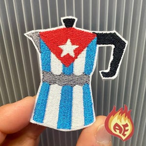 May include: Embroidered patch in the shape of a coffee maker, featuring a red top with a white star, blue and white stripes, and a black handle. A small, flaming logo with the letters "AE" is also visible.