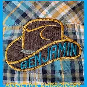 May include: A brown cowboy hat with a yellow brim and a blue embroidered name "BENJAMIN" on the front. The hat is on a blue and yellow plaid fabric.