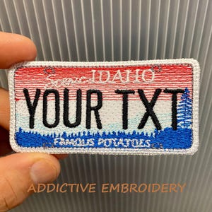 May include: Embroidered patch resembling an Idaho license plate. The patch features red and white stripes at the top, with the word "IDAHO" and the text "YOUR TXT" in black. The bottom has blue and white details with the words "FAMOUS POTATOES".