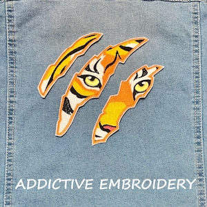 Op de afbeelding: Geborduurde tijgerklauwpatches in oranje, geel, zwart en wit, aangebracht op een lichtblauwe spijkerjas. De patches hebben gedetailleerde oogontwerpen. De tekst "ADDICTIVE EMBROIDERY" staat onderaan.