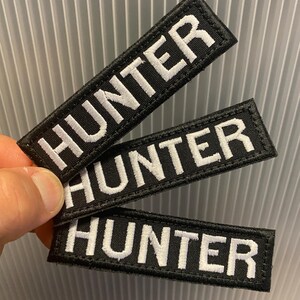 Puede incluir: Tres parches rectangulares negros con la palabra "HUNTER" bordada en blanco. Los parches tienen un borde blanco y est&aacute;n sujetos con una mano. El fondo es una superficie gris claro y acanalada.