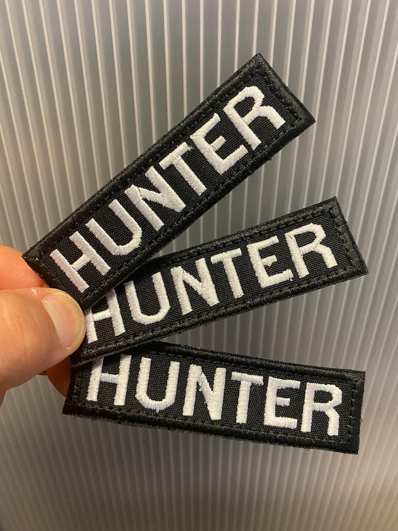Puede incluir: Tres parches rectangulares negros con la palabra "HUNTER" bordada en blanco. Los parches tienen un borde blanco y est&aacute;n sujetos con una mano. El fondo es una superficie gris claro y acanalada.