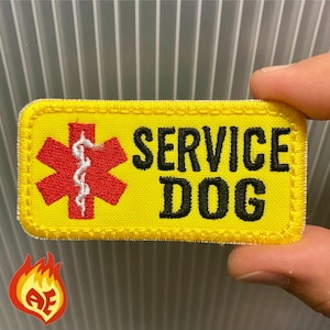 Puede incluir: Un parche rectangular amarillo con texto bordado en negro que dice "SERVICE DOG". El parche presenta una cruz roja con un símbolo de caduceo blanco. El parche tiene un borde amarillo.