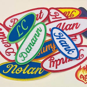 Puede incluir: Una colección de parches bordados de forma ovalada con varios nombres e iniciales, incluidos LC, Danann, Alan, Hank y Nolan. Los parches están representados en diversos colores como azul, blanco, rojo y amarillo con bordes cosidos.