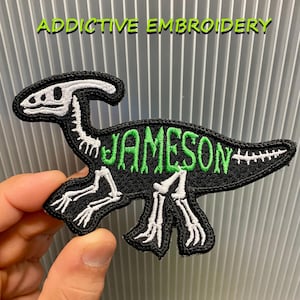 May include: Embroidered patch in the shape of a dinosaur skeleton, with the name "JAMESON" in green lettering on a black background. The patch also has the text "ADDICTIVE EMBROIDERY" in green.