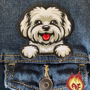Może przedstawiać: Haftowana naszywka z uśmiechniętym białym psem z szarymi akcentami i czerwonym językiem, umieszczona na niebieskiej kurtce dżinsowej. Naszywka ma czarną obwódkę. Widoczna jest również mała naszywka w kształcie płomienia z literami "AE".