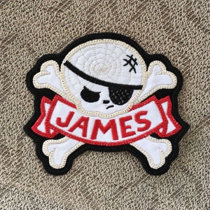 Może przedstawiać: Haftowana naszywka z motywem czaszki i skrzyżowanych piszczeli z przepaską na oku. Imię "JAMES" jest wyświetlane na czerwono-białym banerze. Naszywka ma czarną obwódkę i znajduje się na teksturowanym tle.