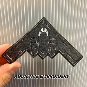 May include: Black embroidered patch in the shape of a stealth bomber. The patch features white stitching and a white emblem. The text "ADDICTIVE EMBROIDERY" is printed in white at the bottom of the image.