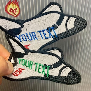 May include: Two embroidered patches in the shape of a space shuttle. The patches are white with black and blue accents, and the text "YOUR TEXT" is embroidered in blue and green. The word "USA" is embroidered in red.