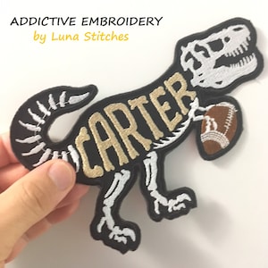 May include: Embroidered patch of a skeleton dinosaur holding a football with the name "CARTER" written across the body. The dinosaur is black with white bones and the name is gold.