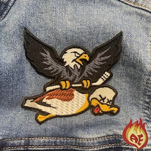 May include: Embroidered patch featuring an eagle with outstretched wings holding a hockey stick, riding a duck. The patch is on a denim jacket. A small, red and yellow flame logo is in the corner.