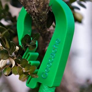 May include: Bright green geocache tool hanging from a tree branch. The tool has the word "Geocache" printed in blue. The tool is made of a smooth, solid material and has a textured handle.