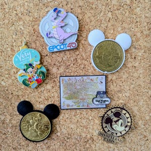 Op de afbeelding: Verzameling Disney-thema pins en een kaart. Inclusief een pin met Figment, een Mickey Mouse-munt, een kaart van Magic Kingdom en een pin met Mickey Mouse. De pins bevatten tekst zoals "EPCOT 40" en "Disneyland Paris".