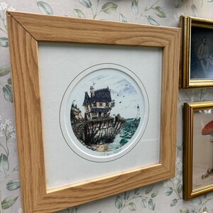 Può includere: Una stampa incorniciata con un'illustrazione circolare di una casa su una scogliera che si affaccia sul mare. L'opera d'arte è incorniciata in legno marrone chiaro. Altre immagini incorniciate sono visibili sullo sfondo, con una carta da parati floreale.