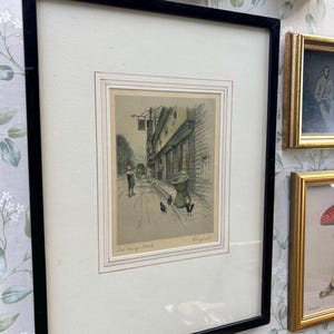May include: Framed print depicting a street scene with a pub sign, labelled "The King's Head." The artwork is an illustration in muted tones, showing figures and buildings. The frame is black.