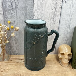 Può includere: Una brocca in ceramica verde scuro con superficie testurizzata, motivi a rami floreali in rilievo e interno bianco. Il recipiente cilindrico presenta un manico curvo e una fascia decorativa attorno al bordo con una finitura opaca.