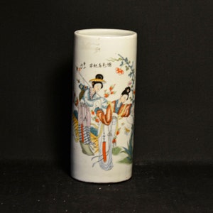 May include: White porcelain vase with a cylindrical shape. The vase is decorated with a hand-painted scene of two women in traditional Chinese clothing, surrounded by flowers and foliage. The vase has Chinese characters written on it.
