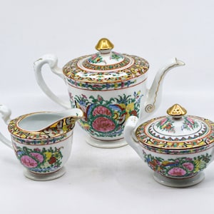 May include: A porcelain tea set featuring a teapot, creamer, and sugar bowl. The set is white with colorful floral and bird designs, gold accents, and a gold-colored finial on the teapot and sugar bowl lid.
