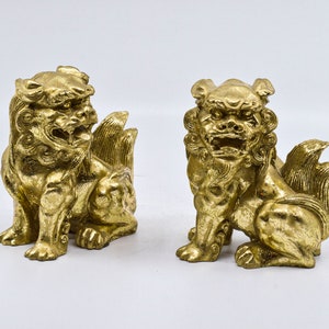 May include: Two gold-colored, decorative lion statues with intricate details. The statues are sitting on a white surface.