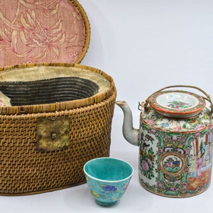 May include: A wicker basket with a lid, a porcelain teapot with a floral design, and a small blue teacup. The teapot has a handle and a lid with a floral design. The teacup has a floral design on the inside.