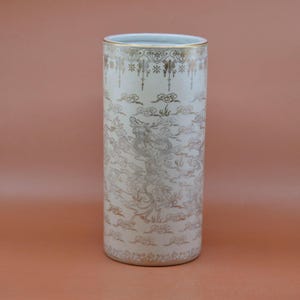 May include: A white ceramic vase with a gold floral design. The vase has a cylindrical shape and is decorated with a pattern of flowers and leaves.