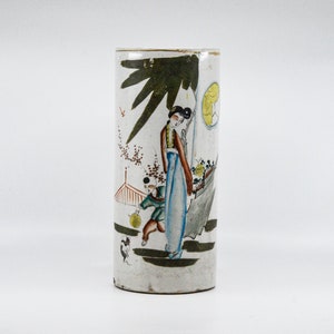 May include: A white ceramic cylinder vase with a hand-painted scene of a woman and a child in a garden setting. The scene is painted in shades of green, blue, red, and yellow.