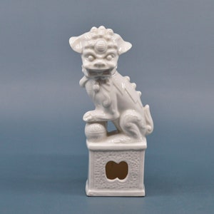 May include: White porcelain figurine of a foo dog, a mythical creature often used as a guardian in Chinese culture. The foo dog is sitting on a square base with a decorative design.