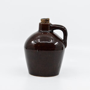 May include: A small brown ceramic jug with a cork stopper and a handle. The jug is glazed and has a rustic look.