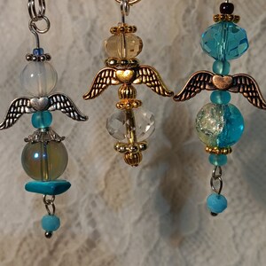 May include: Three angel-shaped charms with wings, each with a different colored glass bead. The first charm has a clear glass bead with gold accents, the second charm has a light blue glass bead with gold accents, and the third charm has a clear glass bead with blue and gold accents.