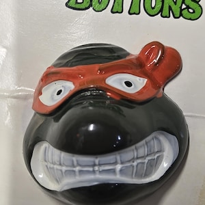 May include: A black and red Teenage Mutant Ninja Turtles button with a white toothy grin. The button features a black mask with white eyes and a red bandana.