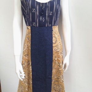 Op de afbeelding: Een mouwloze jurk met een ronde hals. De lijfjes hebben een marineblauw en wit gestreept patroon. De rok heeft een centraal marineblauw paneel geflankeerd door panelen met een paisley-print in tinten goud, bruin en crème. Donkere knoopdetail bij de halslijn.