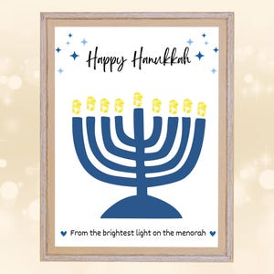 May include: A framed print with a light wood frame. The print features a blue menorah with yellow flames, the words "Happy Hanukkah" and the phrase "From the brightest light on the menorah" in black script, and blue star accents.