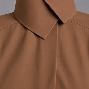 May include: A solid brown shirt featuring a high, structured collar with a sharp, overlapping point design. The garment has a smooth, matte texture and clean, minimalist lines, with visible stitching along the shoulder seams and a concealed placket.