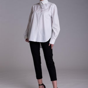 May include: A white long-sleeved button-down shirt with a high collar and a textured white floral appliqu&eacute; detail on the right side, paired with slim-fit black trousers and black high-heeled sandals with ankle straps.