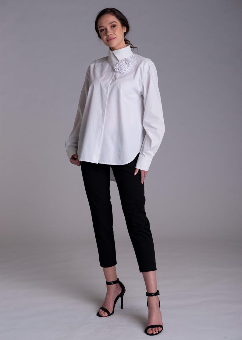 May include: A white long-sleeved button-down shirt with a high collar and a textured white floral appliqu&eacute; detail on the right side, paired with slim-fit black trousers and black high-heeled sandals with ankle straps.