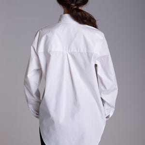 May include: A crisp white long-sleeved button-down shirt featuring a classic collar, a central back box pleat, and a curved hemline. The shirt is worn over plain black trousers, showcasing a clean, structured silhouette with long sleeves.