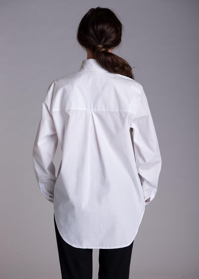 May include: A crisp white long-sleeved button-down shirt featuring a classic collar, a central back box pleat, and a curved hemline. The shirt is worn over plain black trousers, showcasing a clean, structured silhouette with long sleeves.