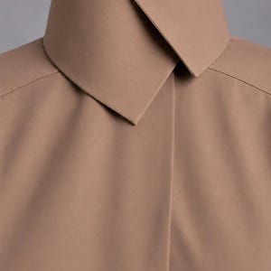May include: A solid tan shirt featuring a high, structured collar with a sharp, overlapping point design. The garment has a smooth, matte finish and clean, minimalist lines, focusing on the tailored construction of the neckline and shoulder seams.