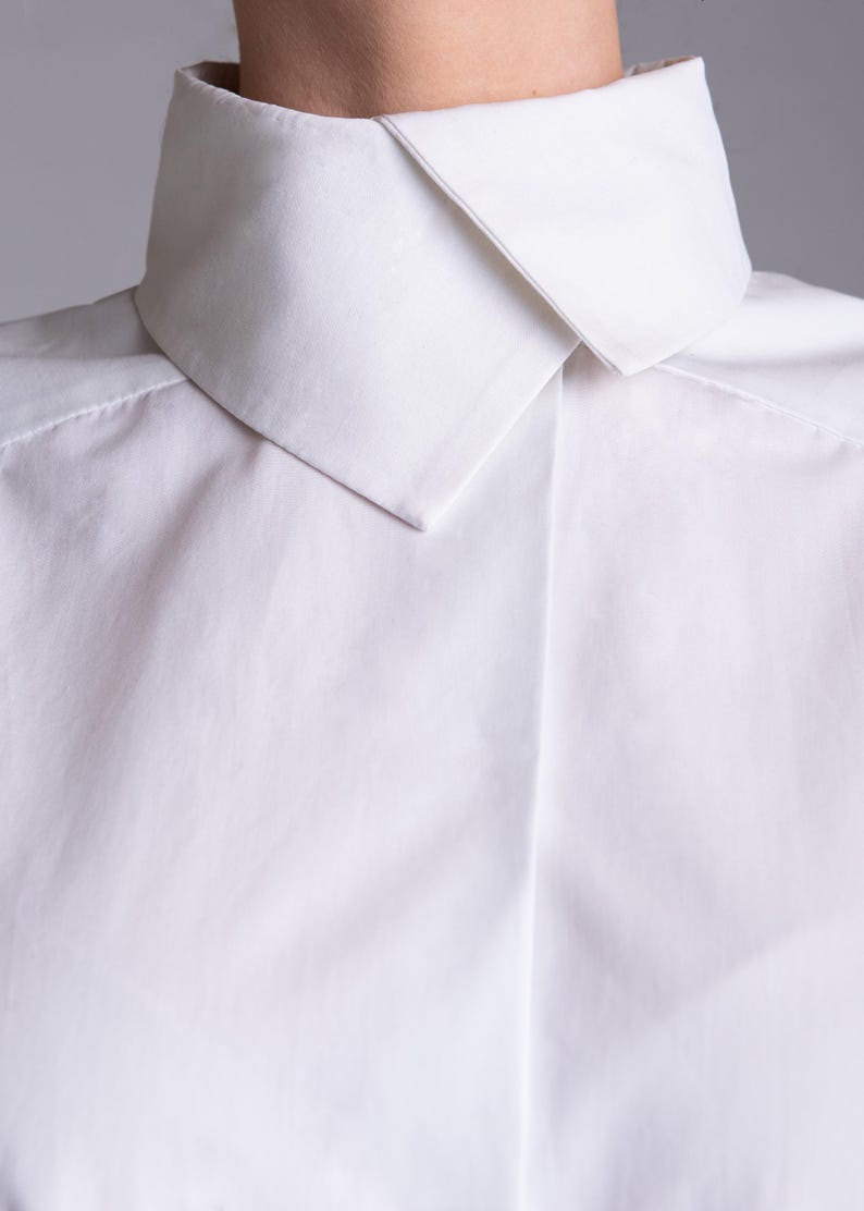 May include: A crisp white button-down shirt featuring a structured high collar with a pointed fold. The garment is made of a smooth, opaque fabric with a clean, minimalist design, showcasing a concealed placket and sharp, tailored shoulder seams.