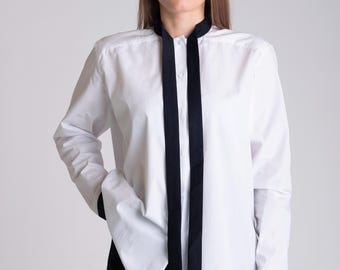 May include: A white long-sleeve shirt with black trim on the collar and cuffs. Two black fabric strips hang vertically down the front of the shirt. The shirt is buttoned up the front.