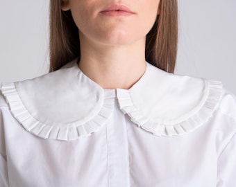 May include: A crisp white button-down shirt featuring a large, rounded Peter Pan collar. The collar is detailed with a delicate, evenly spaced pleated trim along the outer edge, adding texture to the smooth, solid white fabric of the garment.