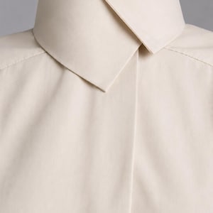 May include: A light beige button-down shirt featuring a high, structured collar with a sharp, pointed fold. The garment has a clean, smooth texture and a concealed placket design, focusing on the neckline and upper chest area of the shirt.
