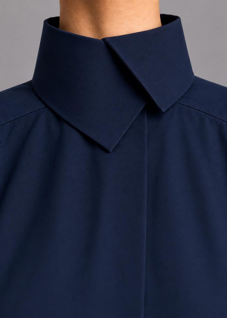 May include: A dark navy blue shirt featuring a high, structured stand-up collar with a pointed, overlapping front closure. The garment is crafted from a smooth, solid-colored fabric with clean, minimalist lines and visible shoulder seam stitching.
