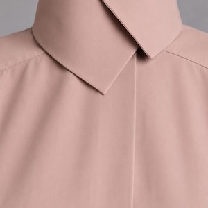 May include: A dusty rose shirt featuring a high, structured collar with a pointed, overlapping design. The garment has a smooth, matte finish and a clean, minimalist front placket that conceals the buttons, creating a seamless appearance.
