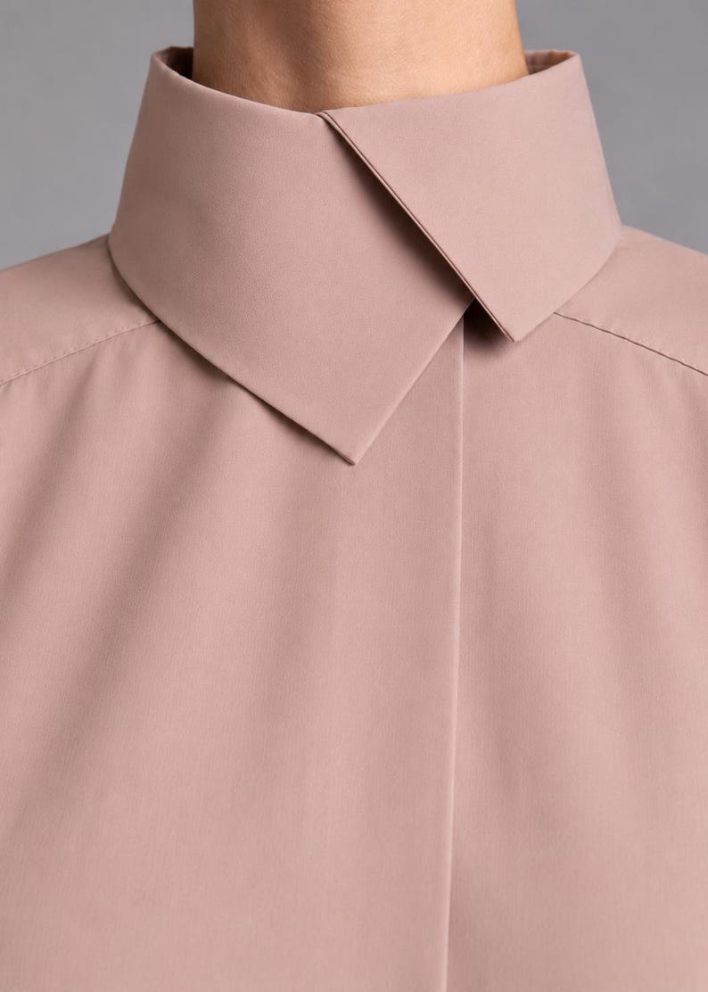 May include: A dusty rose shirt featuring a high, structured collar with a pointed, overlapping design. The garment has a smooth, matte finish and a clean, minimalist front placket that conceals the buttons, creating a seamless appearance.