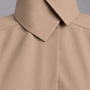 May include: A beige collared shirt featuring a high, structured stand-up collar with a pointed fold-over detail. The garment has a smooth, matte finish and a concealed front placket, showcasing clean lines and a minimalist construction throughout.