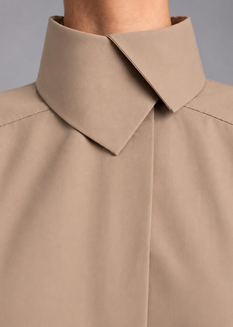 May include: A beige collared shirt featuring a high, structured stand-up collar with a pointed fold-over detail. The garment has a smooth, matte finish and a concealed front placket, showcasing clean lines and a minimalist construction throughout.