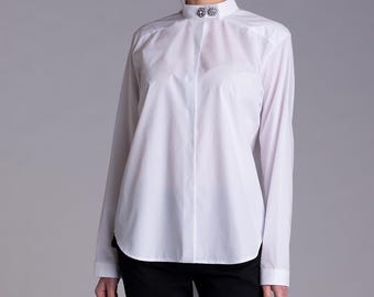 May include: A crisp white long-sleeved button-down shirt featuring a high mandarin collar adorned with two decorative circular silver-toned buttons with embedded clear crystals. The shirt has a clean, minimalist design with a hidden button placket.